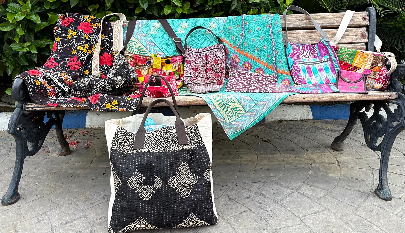 Handmade quilts, totes and bags stitched from sari fabric, laid out on a garden bench.