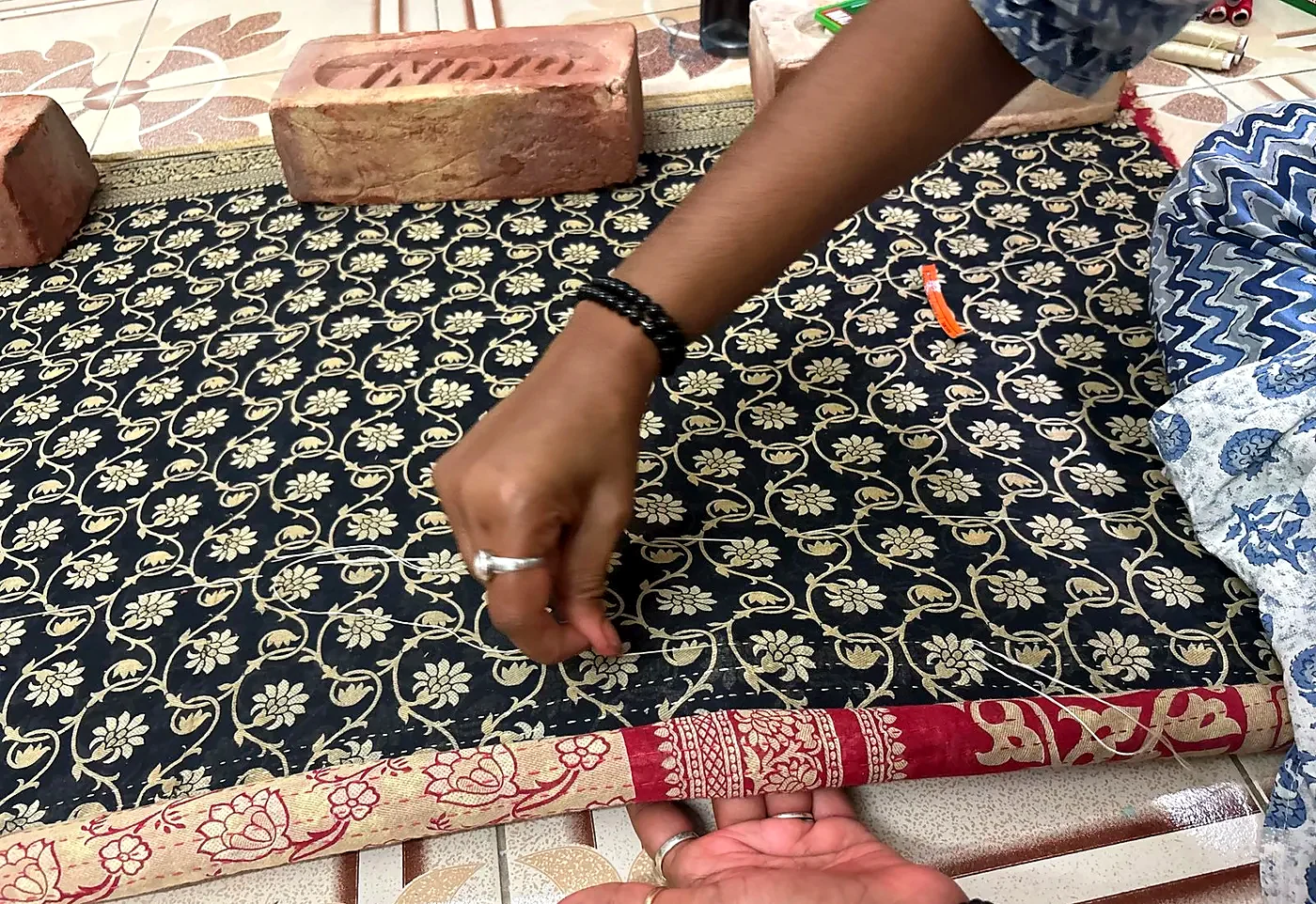 A woman's hands quilting a black and gold block-printed sari, weighted down with bricks.